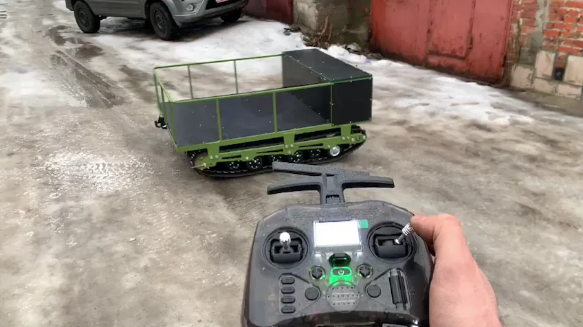 Пульт радиоуправления в руке и гусеничная платформа с кузовом на заснеженной площадке